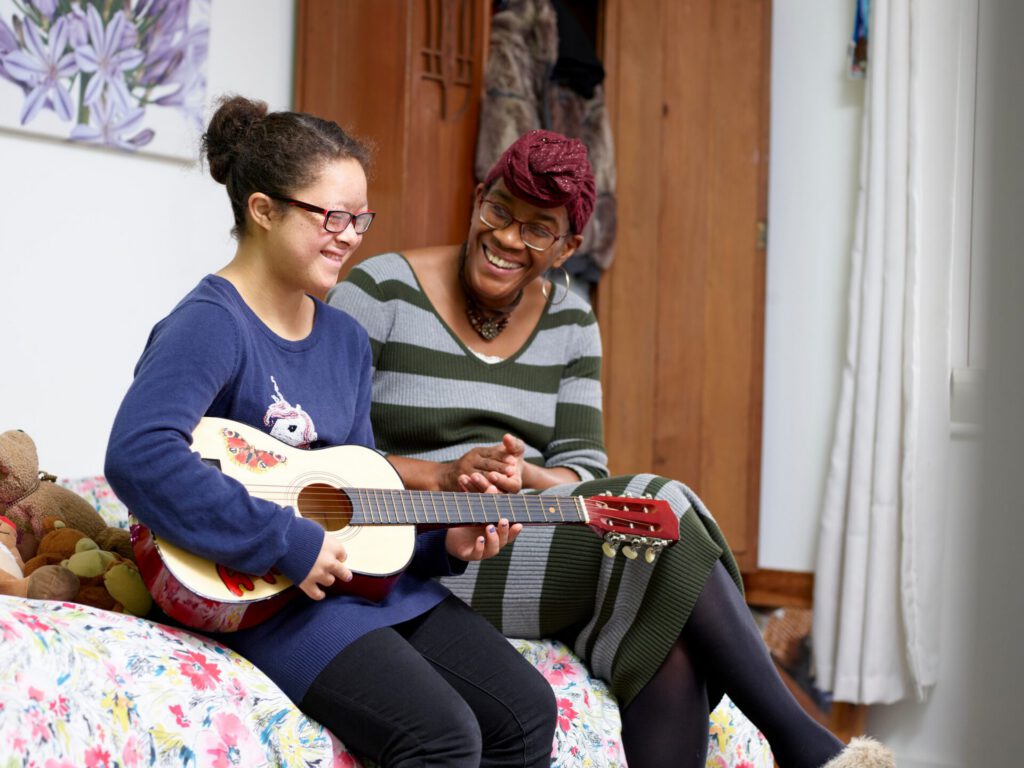 A teenage girl with Down syndrome sits on her bed holding a guitar with her instructor smiling at her progress. 