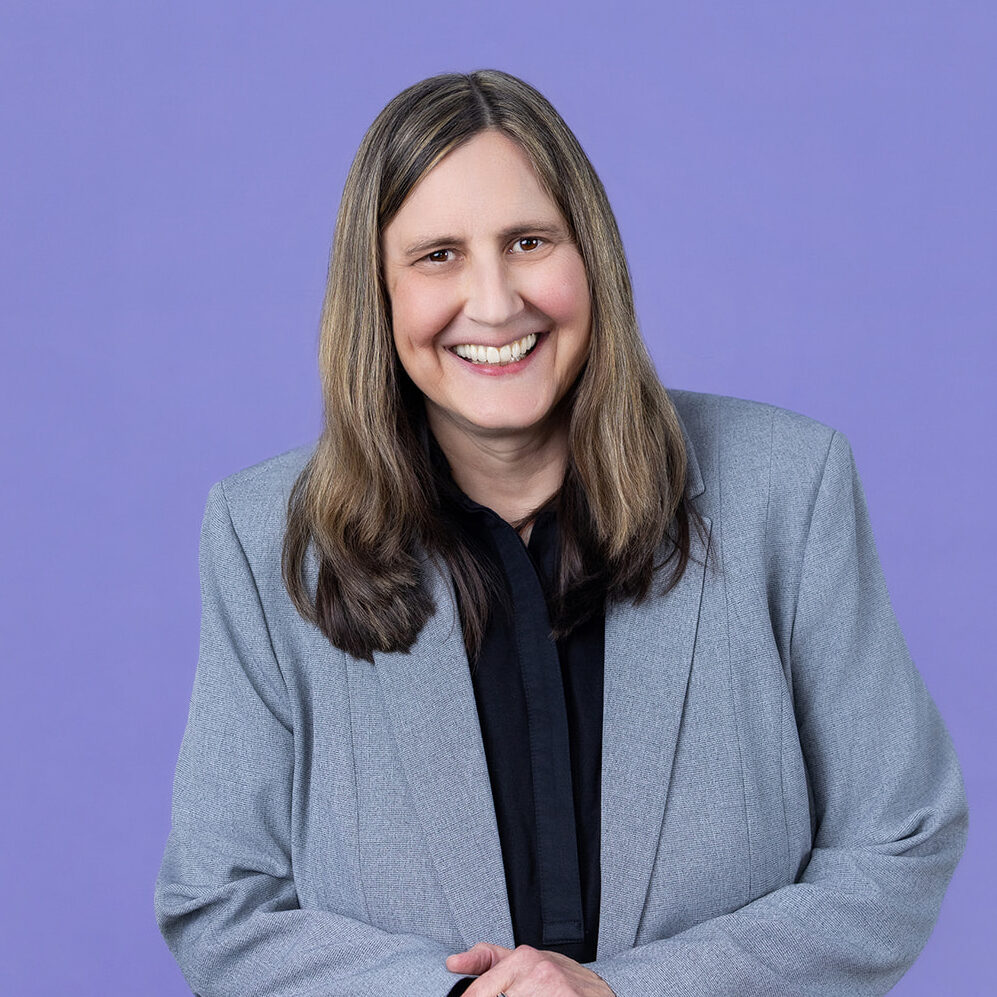 A person with long straight brown hair wearing a light gray blazer over a black shirt stands in front of a light purple background, resting both hands on the back of a black mesh chair and smiling.