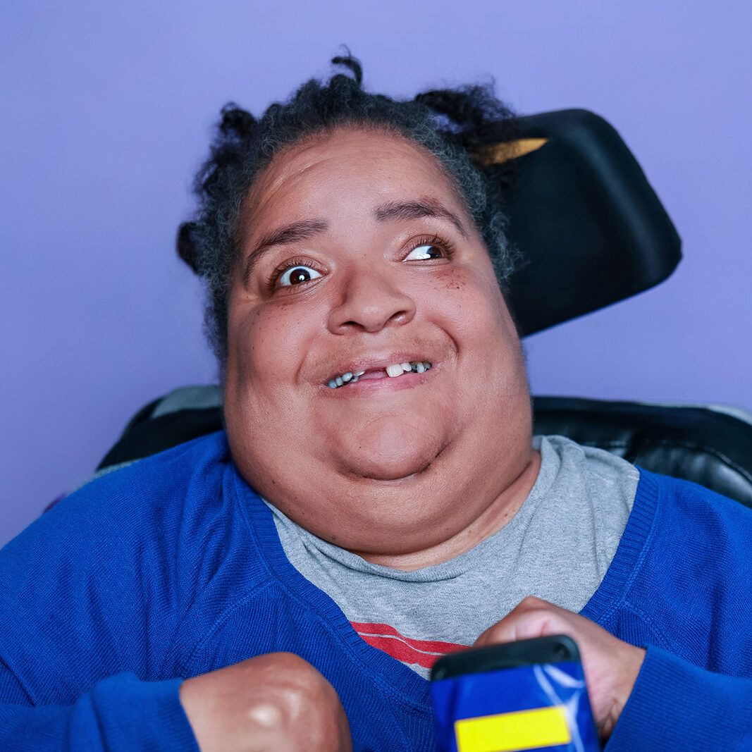 A person with short curly hair wearing a blue top over a gray shirt sits in a wheelchair against a light purple background. The wheelchair has black and blue components, including a control device with yellow stripes.