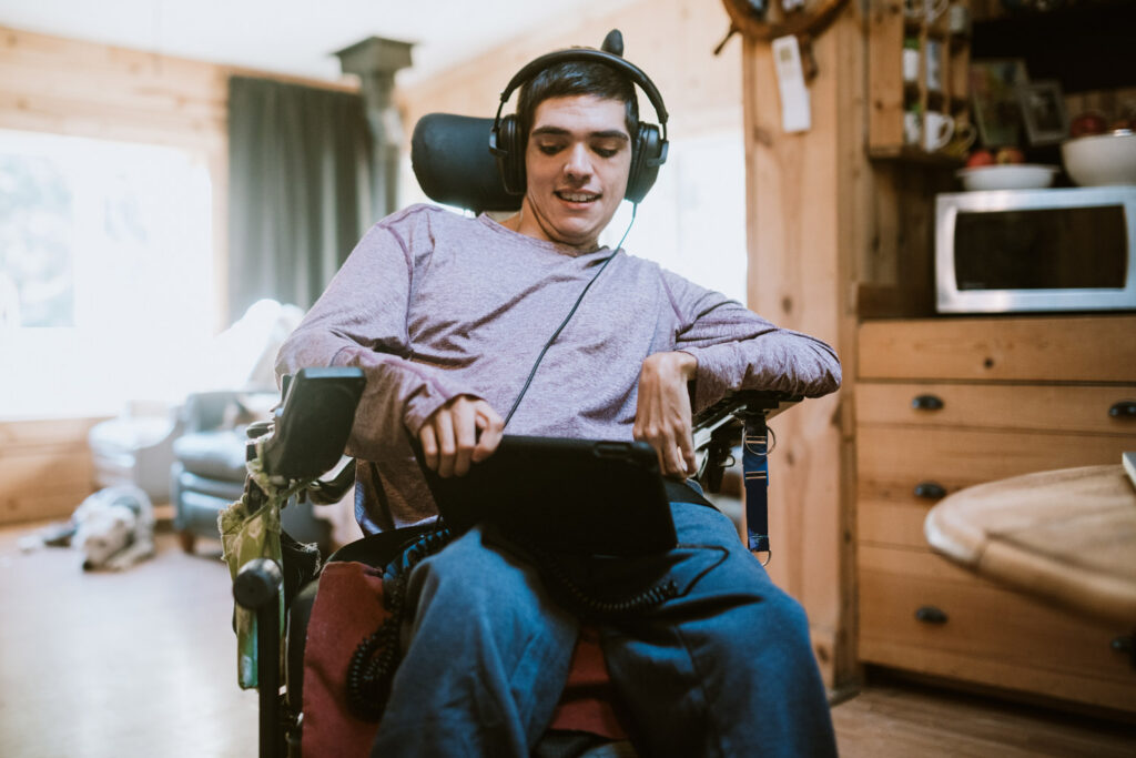 Confident young man in a wheelchair at home using an iPad and listening to a video with headphones on. 