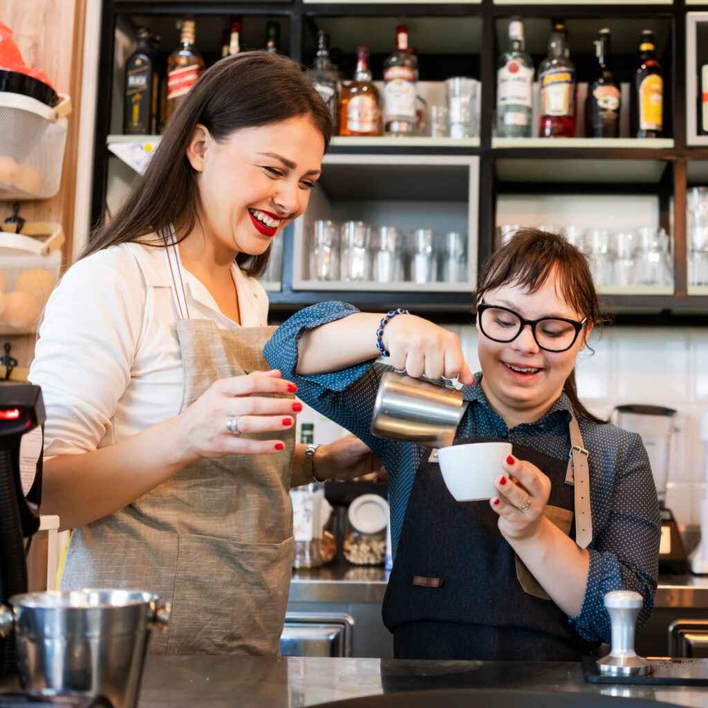 Young woman with Down Syndrome working at cafe pouring coffee. Her coworker stands beside her smiling.
