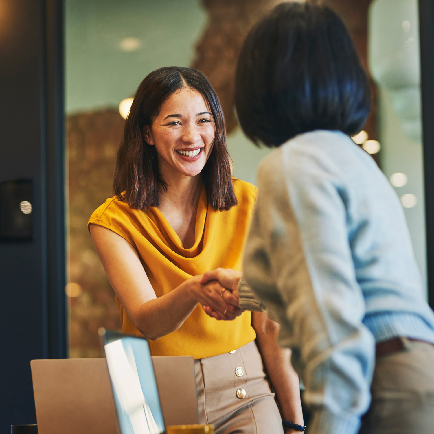Cheerful professional woman in a yellow blouse shaking hands with another person in a meeting room.