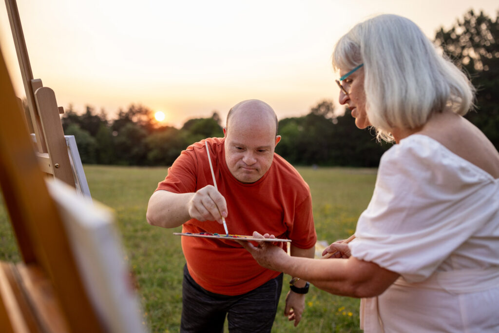 Two people, man with Down syndrome and woman artist painting together outdoors on a meadow.
