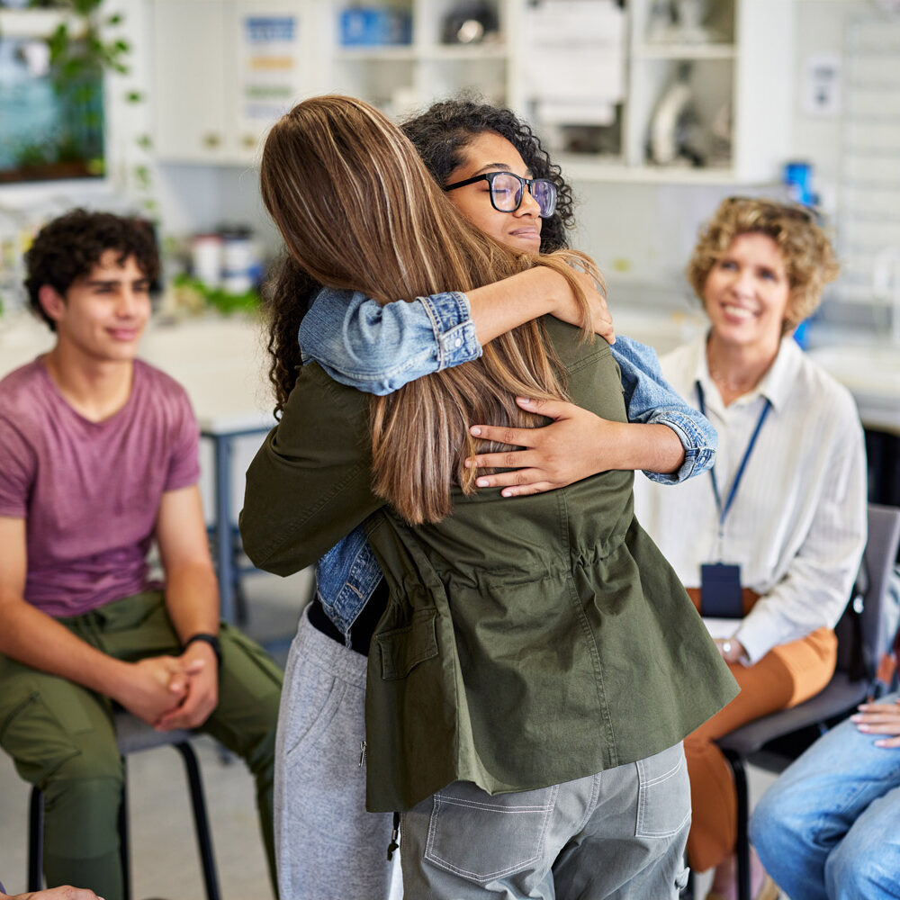 Two female students embracing during a group counseling session, offering comfort and emotional connection. Multiethnic classmates observe moment of support with attentive and compassionate expressions. Therapist smiling in the background, witnessing positive outcome of the session.