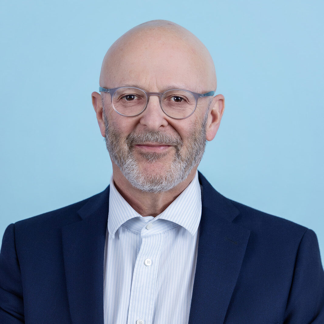 A person with a bald head, a gray beard, and clear framed glasses is wearing a navy blue suit jacket and a white striped dress shirt, standing in front of a light blue background.