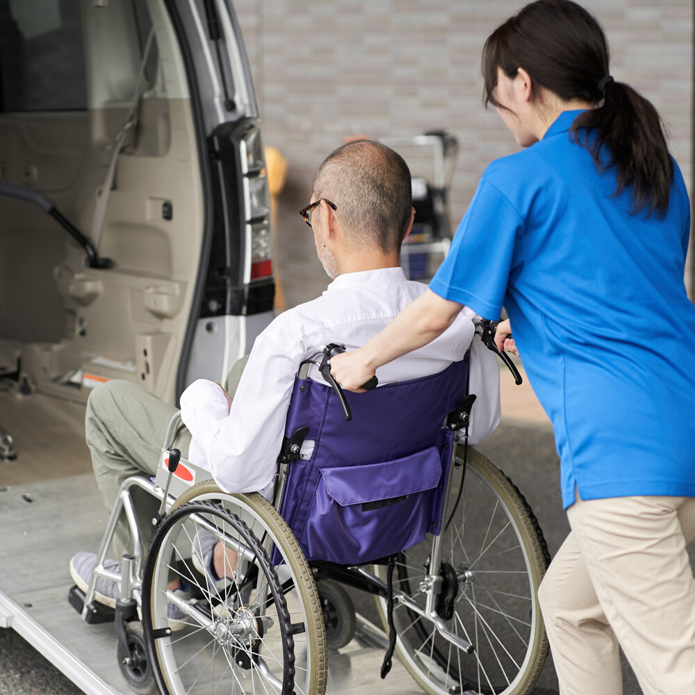 A caregiver wheeling an elderly person in a wheelchair into a taxi.