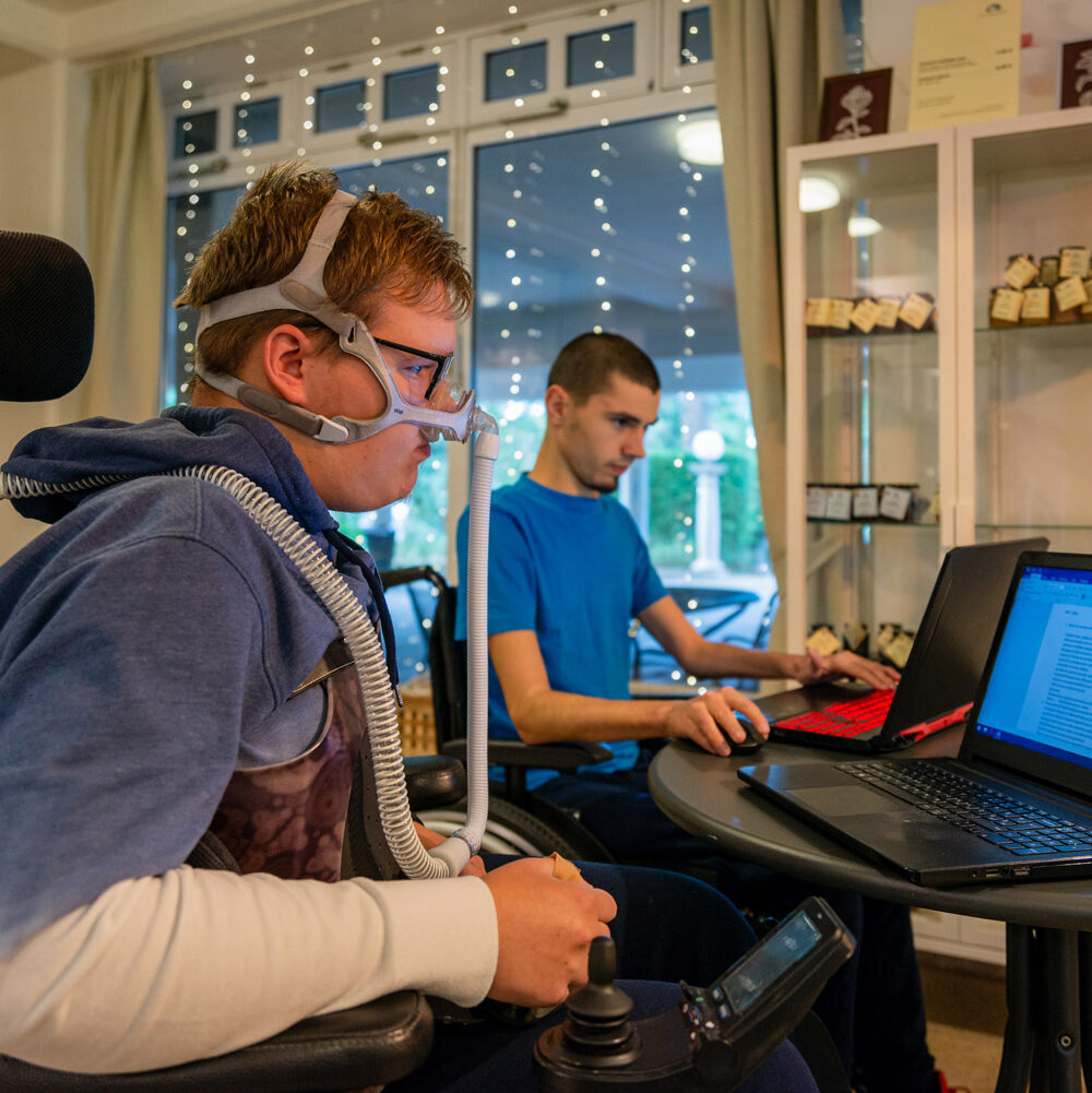 Teenager with dystrophy working on a laptop next to their roommate with twinkling holiday lights in the background.