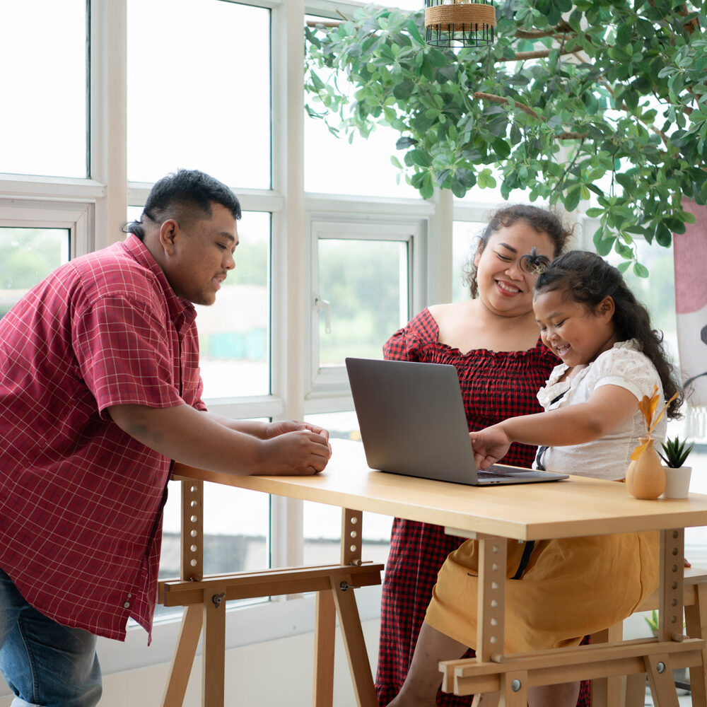 Hispanic family with a father wearing a prosthetic leg, is happily assisting a child as she uses a laptop. Behind them is a bright, office building with large windows.