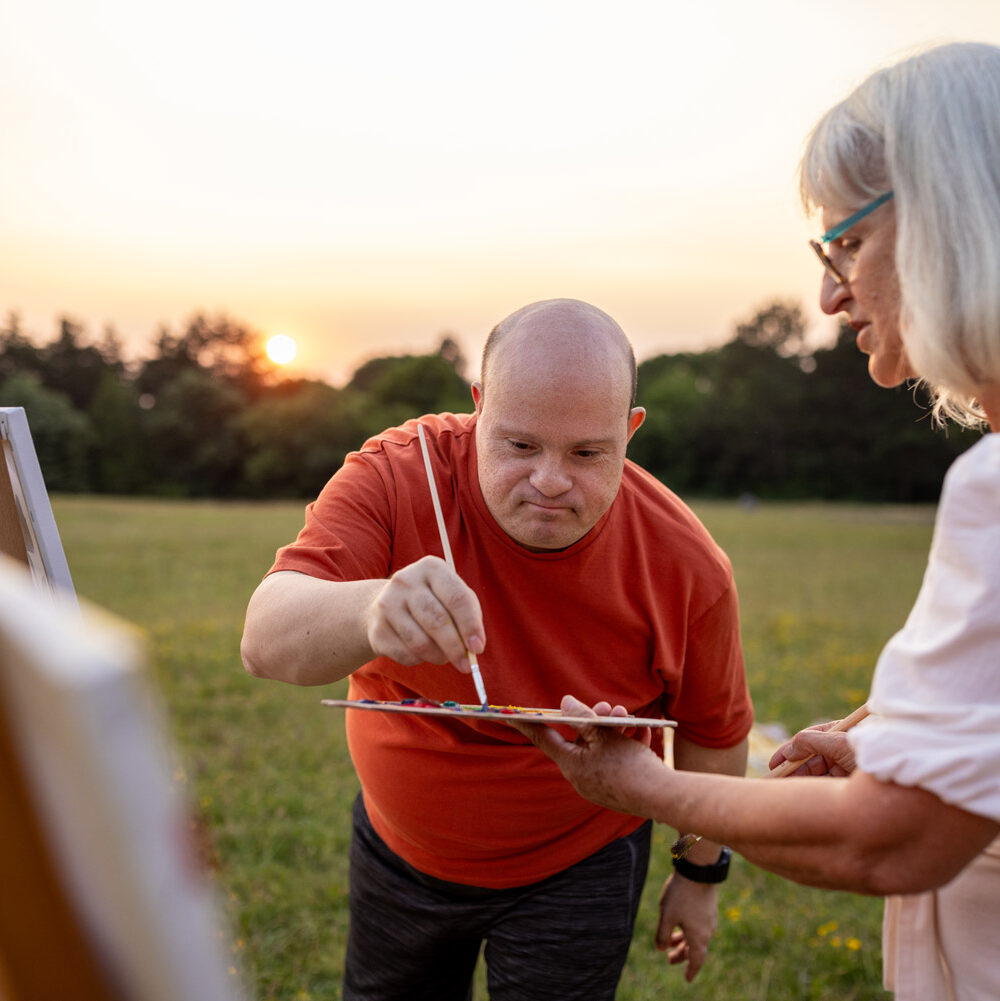 Two people, man with Down syndrome and woman artist painting together outdoors on a meadow.