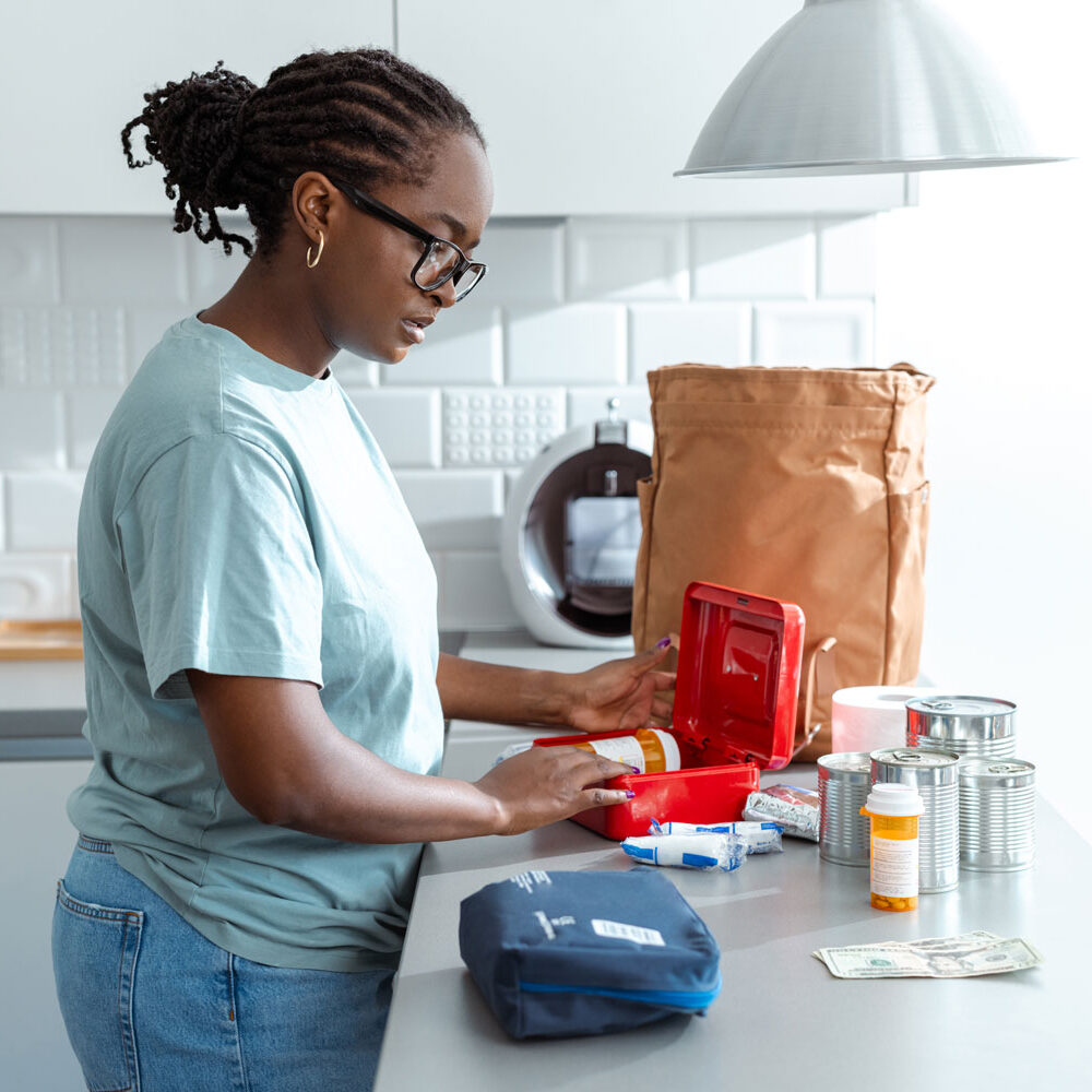 A young, Black woman organizes a first aid kit at the kitchen counter, placing supplies like medications and bandages into the red medical box, with a paper bag and cans nearby.