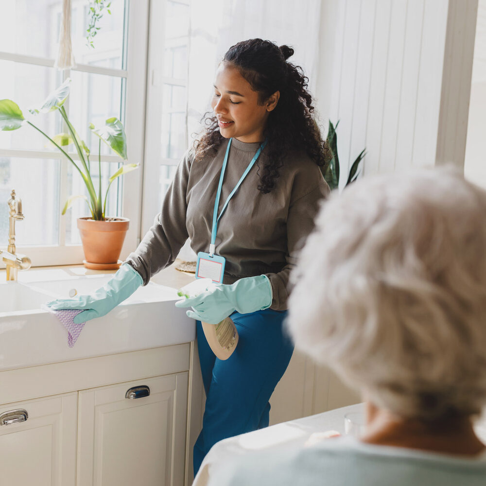 Black woman with a name tag teaching an adult woman how to clean kitchen carefully using spray and microfiber cloth.